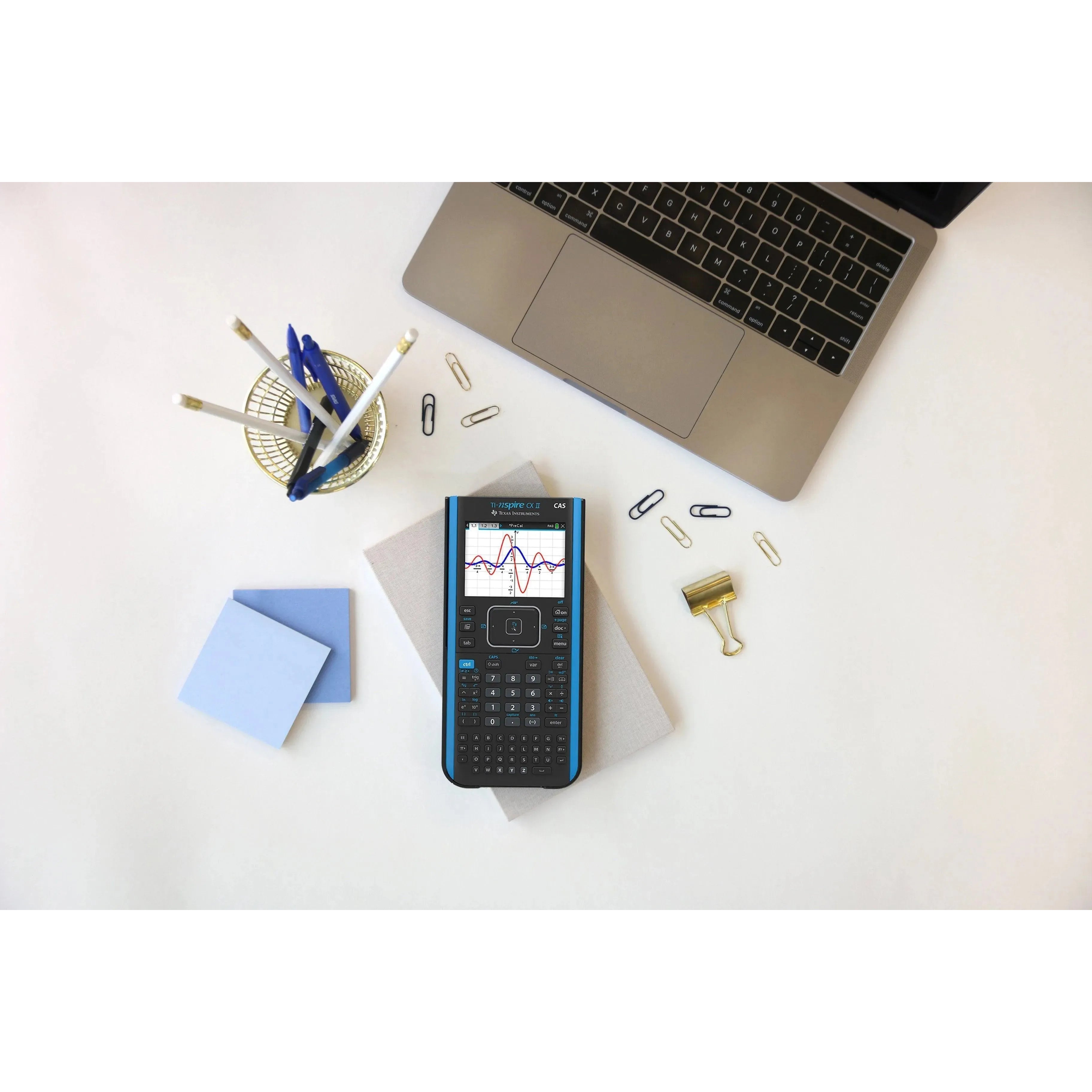 TI-Nspire CX II CAS graphing calculator on desk with laptop, stationery, and paper clips